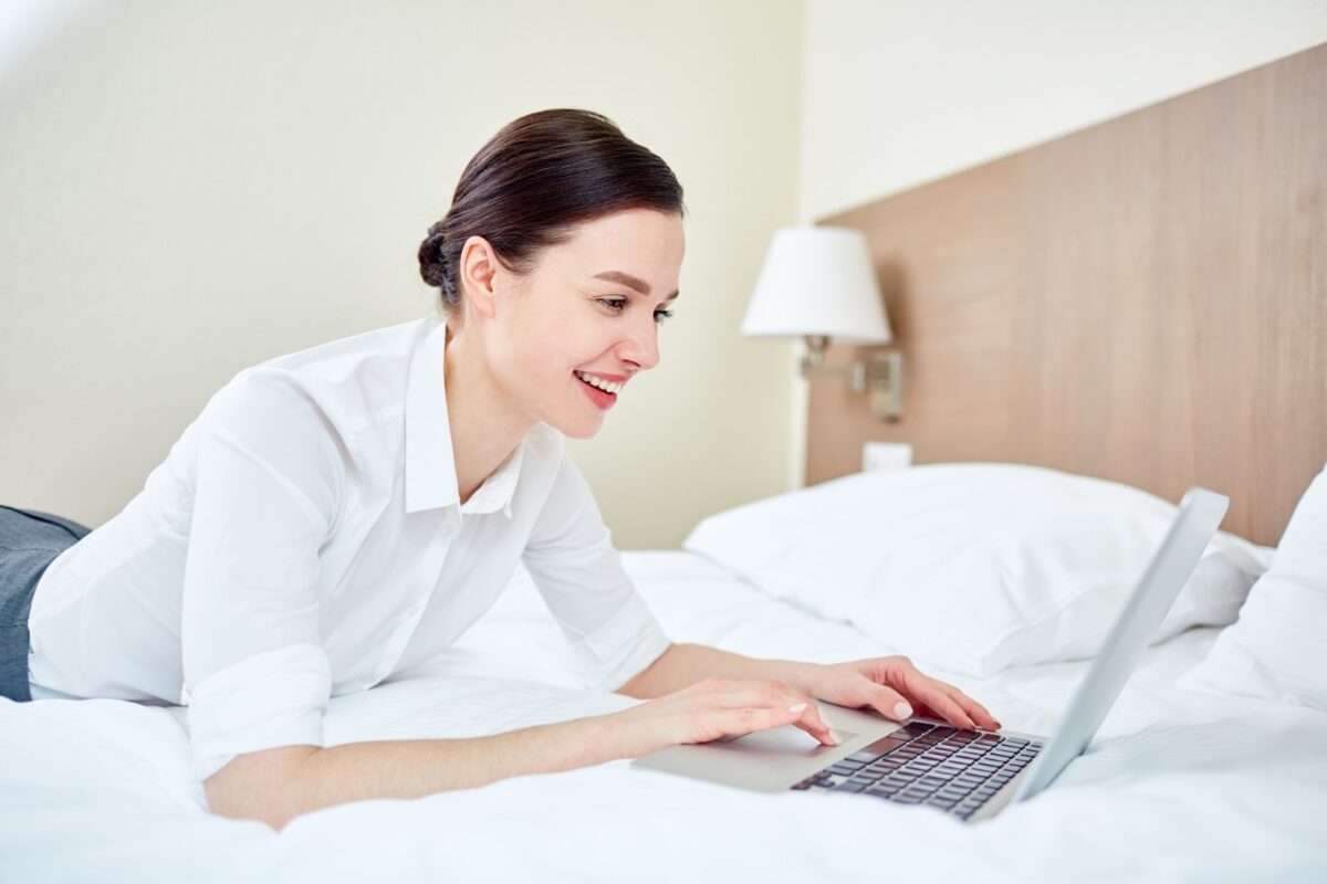 a smiling woman on her hotel bed using a macbook laptop